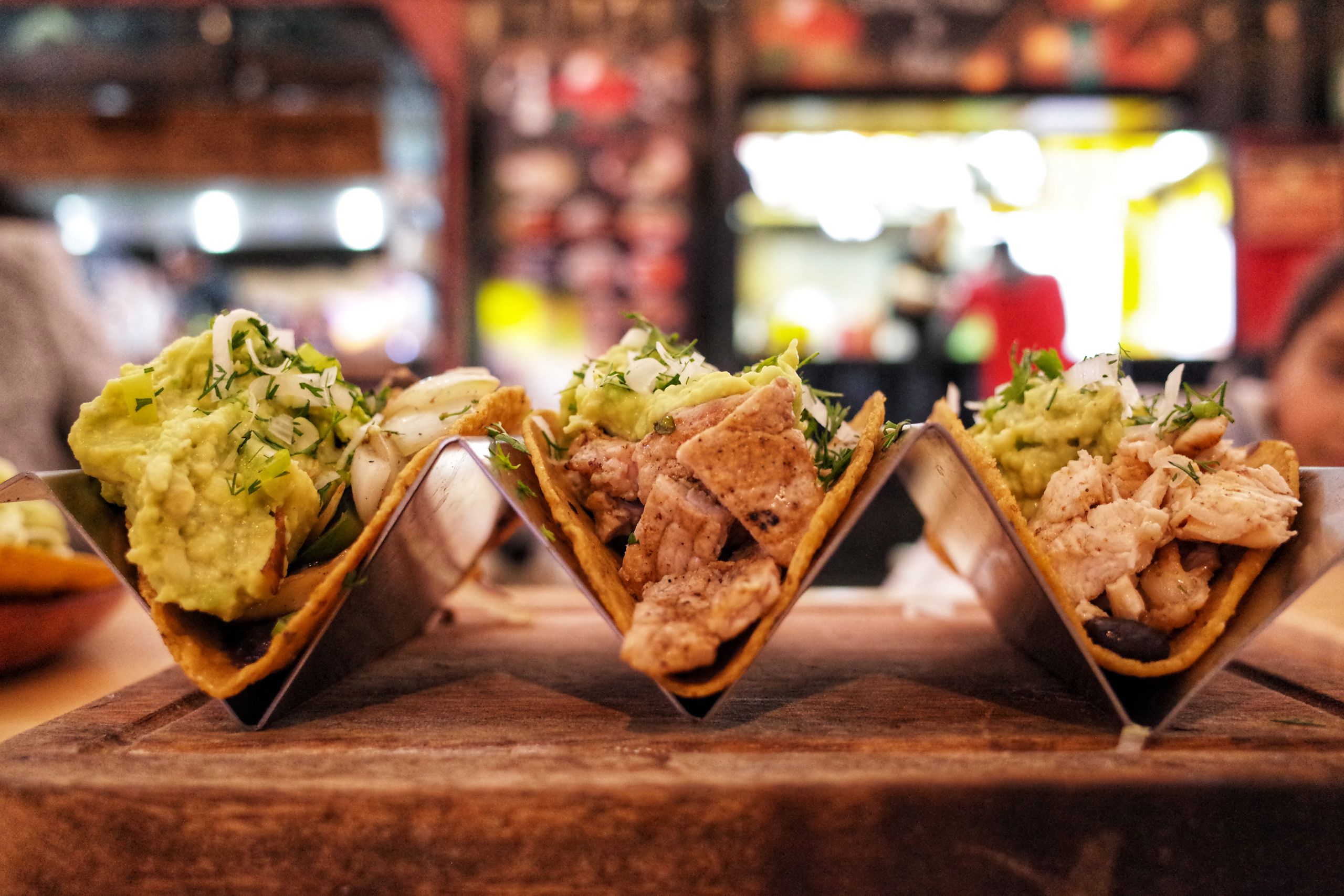 Close-up of three chicken tacos with guacamole, herbs, and onions in a restaurant setting.